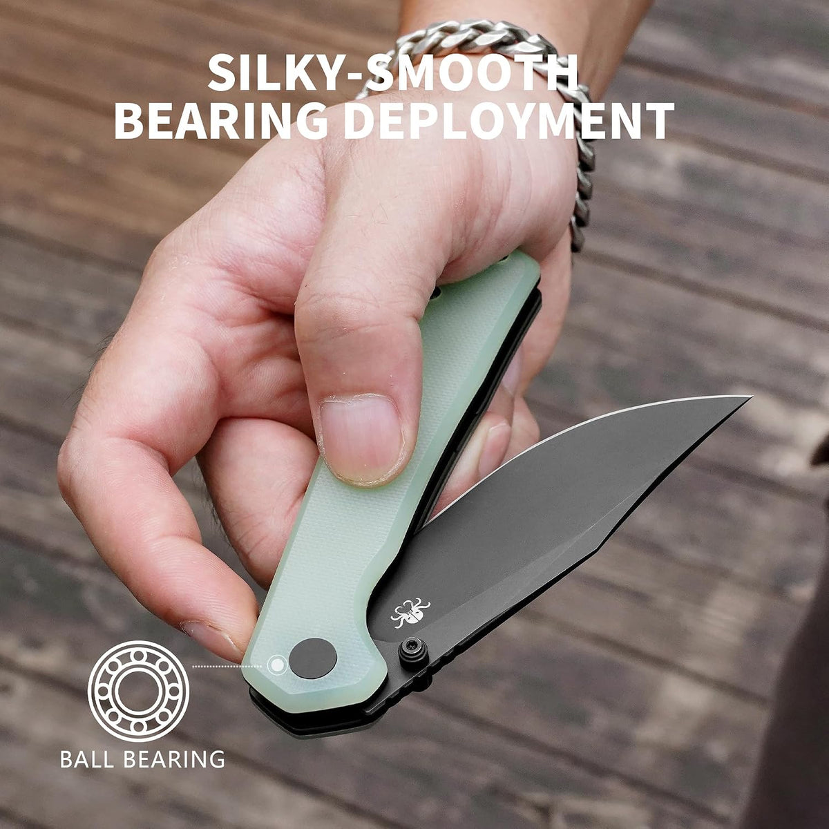 A hand holding an open folding knife with a pale green handle and black blade, over a wooden surface. White text reads 'SILKY-SMOOTH BEARING DEPLOYMENT' and a small diagram says 'BALL BEARING' to explain the product feature.