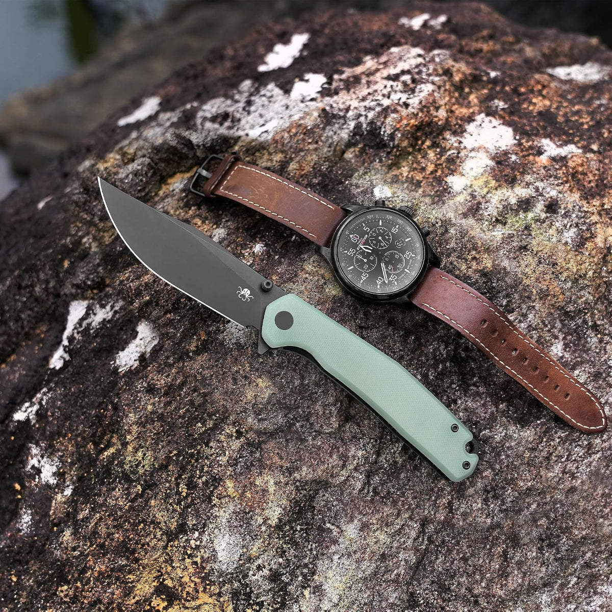 A folding pocket knife with a sage green handle and matte black blade is placed diagonally on a textured brown and white rock, next to a black dial watch with a brown leather strap.