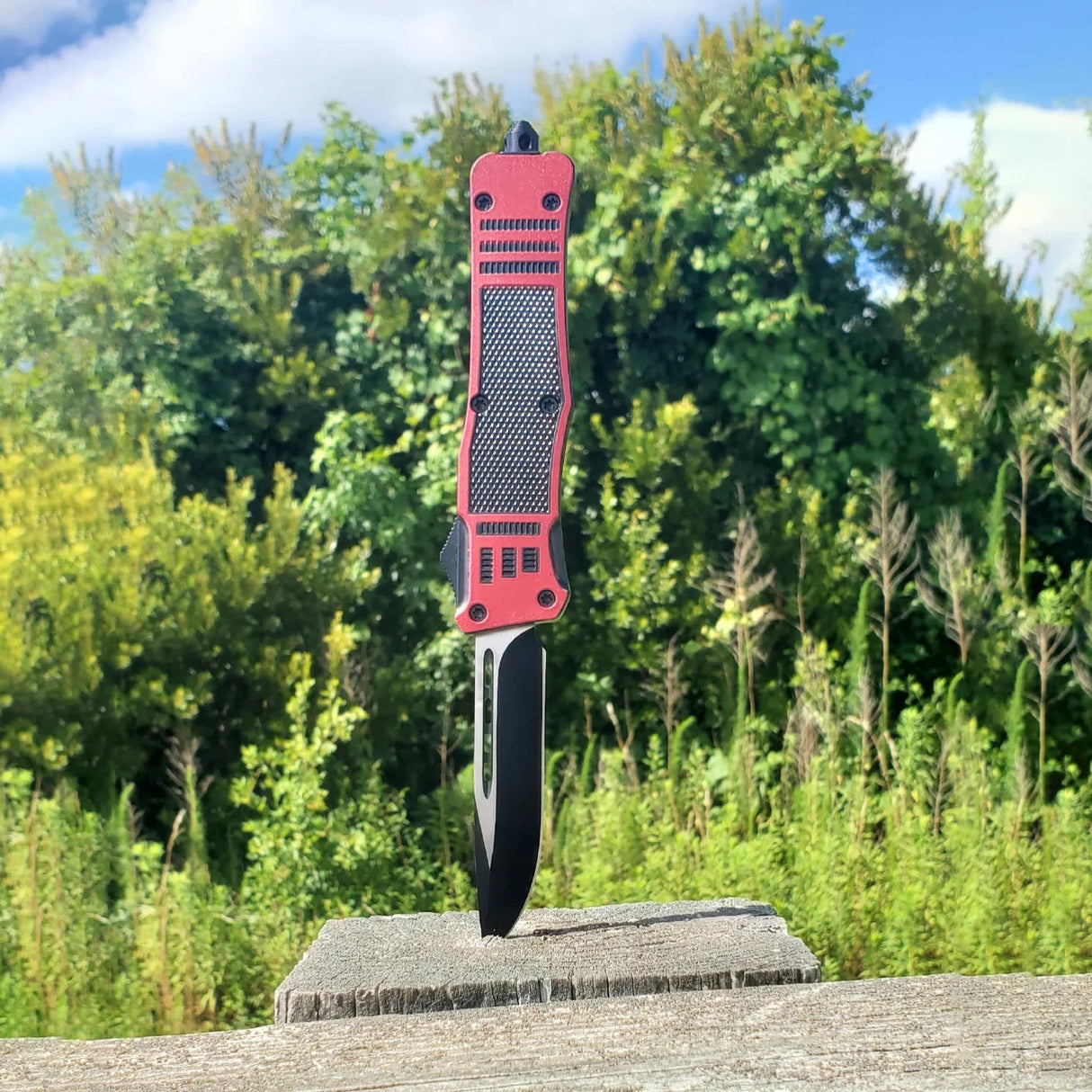 An open black and red 3.25" Armed Force Tactical Automatic OTF Pocket Knife pierced through a wood vertically with greenery background