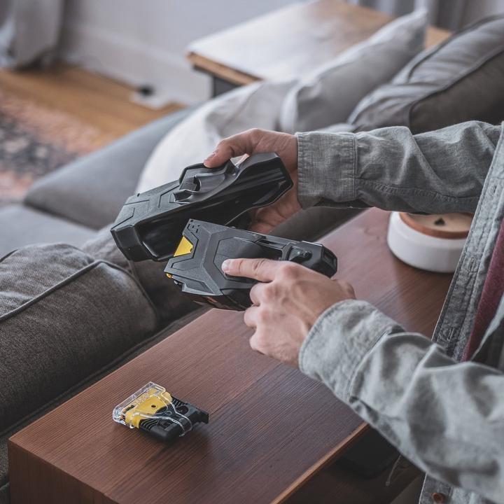 Hands assembling BLACKHAWK! TASER X2 in a living room setting, next to a spare cartridge