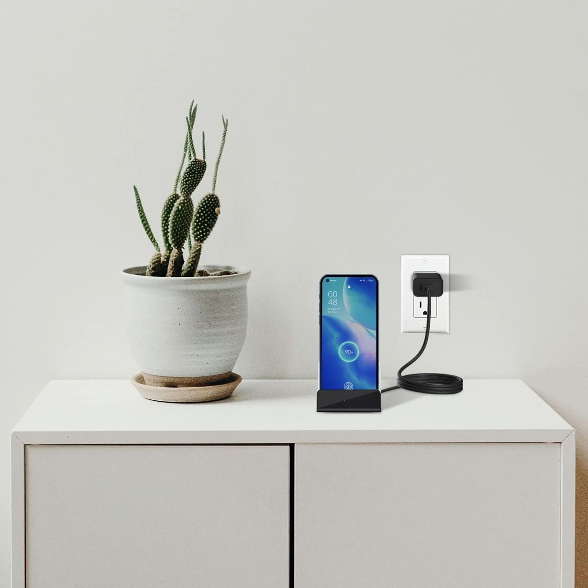 SpyWfi USB-C Phone Charger Hidden Camera disguised as a phone charging dock, sits on a white cabinet next to a plant, plugged into a wall outlet