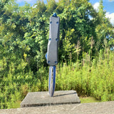 An open blue 3.25" Armed Force Tactical OTF Dagger Knife pierced through a wood vertically with greenery background
