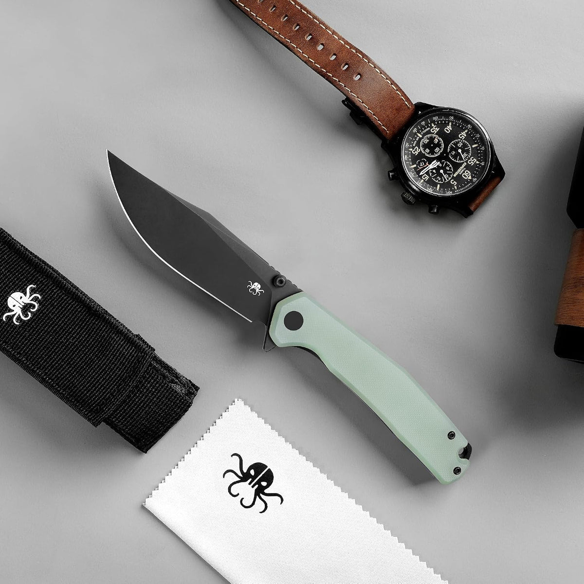 A fixed-blade knife with a matte black blade and a pale mint green handle is arranged on a light gray surface alongside a black nylon sheath, a white microfiber cloth, and a brown leather watch strap with a black chronograph watch.