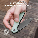 A hand holding an open folding knife with a pale green handle and black blade, over a wooden surface. White text reads 'SILKY-SMOOTH BEARING DEPLOYMENT' and a small diagram says 'BALL BEARING' to explain the product feature.