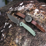 A folding pocket knife with a sage green handle and matte black blade is placed diagonally on a textured brown and white rock, next to a black dial watch with a brown leather strap.